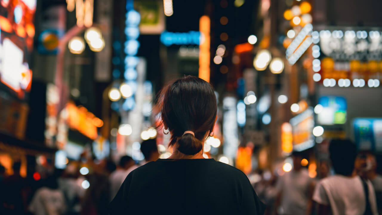A rear view of a woman with her brown hair tied back in a low bun, walking through a crowded city street at night. The camera focus is sharply on her, while the bustling city ahead dissolves into a soft-focus bokeh background of vibrant glowing neon signs, streetlights, and building displays in shades of orange, yellow, and blue.