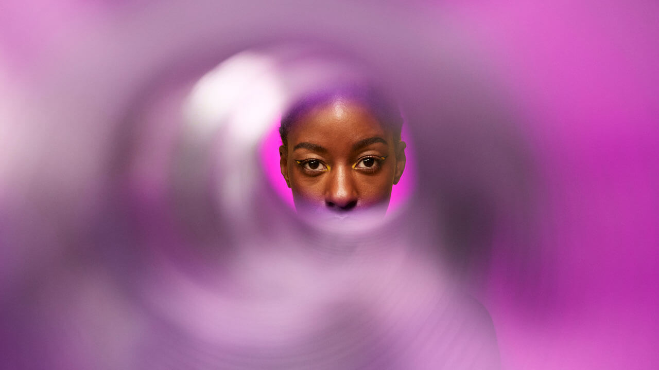 A close-up portrait of a Black woman looking forward, viewed through a blurred circular lens or tube on a bright magenta background, symbolizing the process of focusing on and verifying real human respondents in market research.