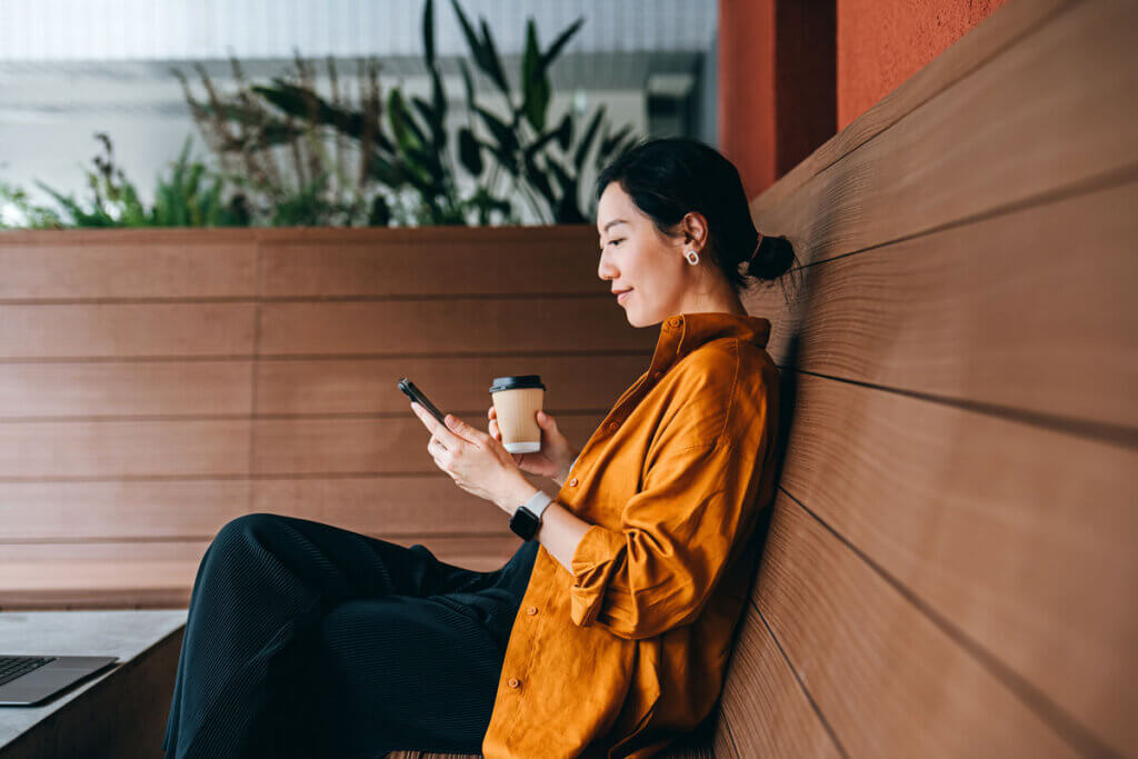 side view of a woman sitting on a wooden bench, looking at her smartphone while holding a coffee cup, representing a consumer participating in a mobile research survey.