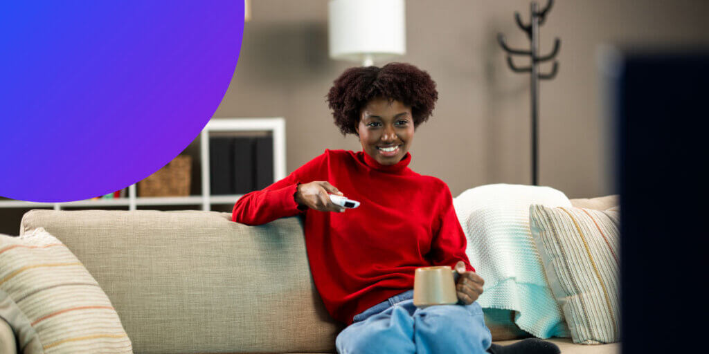 A young Black woman with short curly hair sits comfortably on a beige living room sofa, smiling as she watches television. She wears a bright red turtleneck sweater and jeans, holding a remote control pointed toward the TV in one hand and a coffee mug in the other. A large purple-to-blue gradient circle overlay anchors the top left corner of the frame.