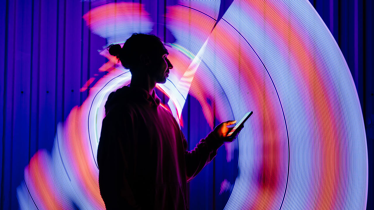 A side-profile silhouette of a person with their hair in a bun holding a smartphone. They are framed by a vibrant, long-exposure light painting background featuring swirling concentric lines of bright red, blue, and white neon light casting a glow against a dark vertical corrugated wall.