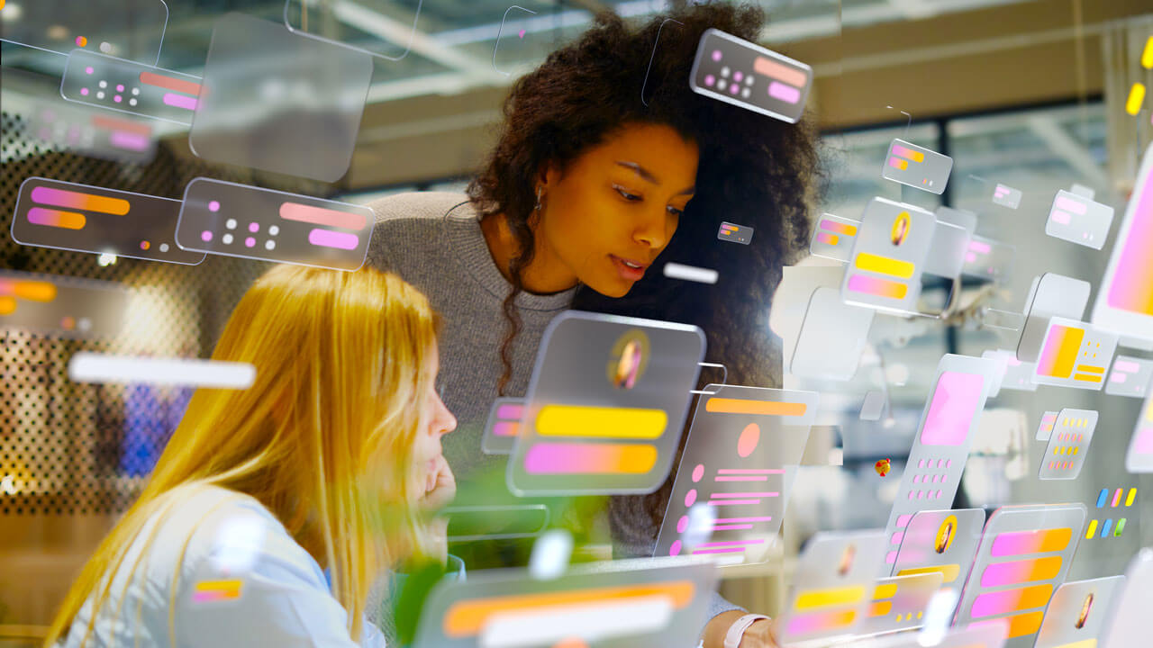 Two female colleagues—one with blonde hair seated and one with curly dark hair leaning in—working together at a desk. They are looking down at a screen, completely overlaid by a futuristic digital graphic layer of floating, translucent user interface windows, text bubbles, and profile panels in shades of pink, yellow, and grey.